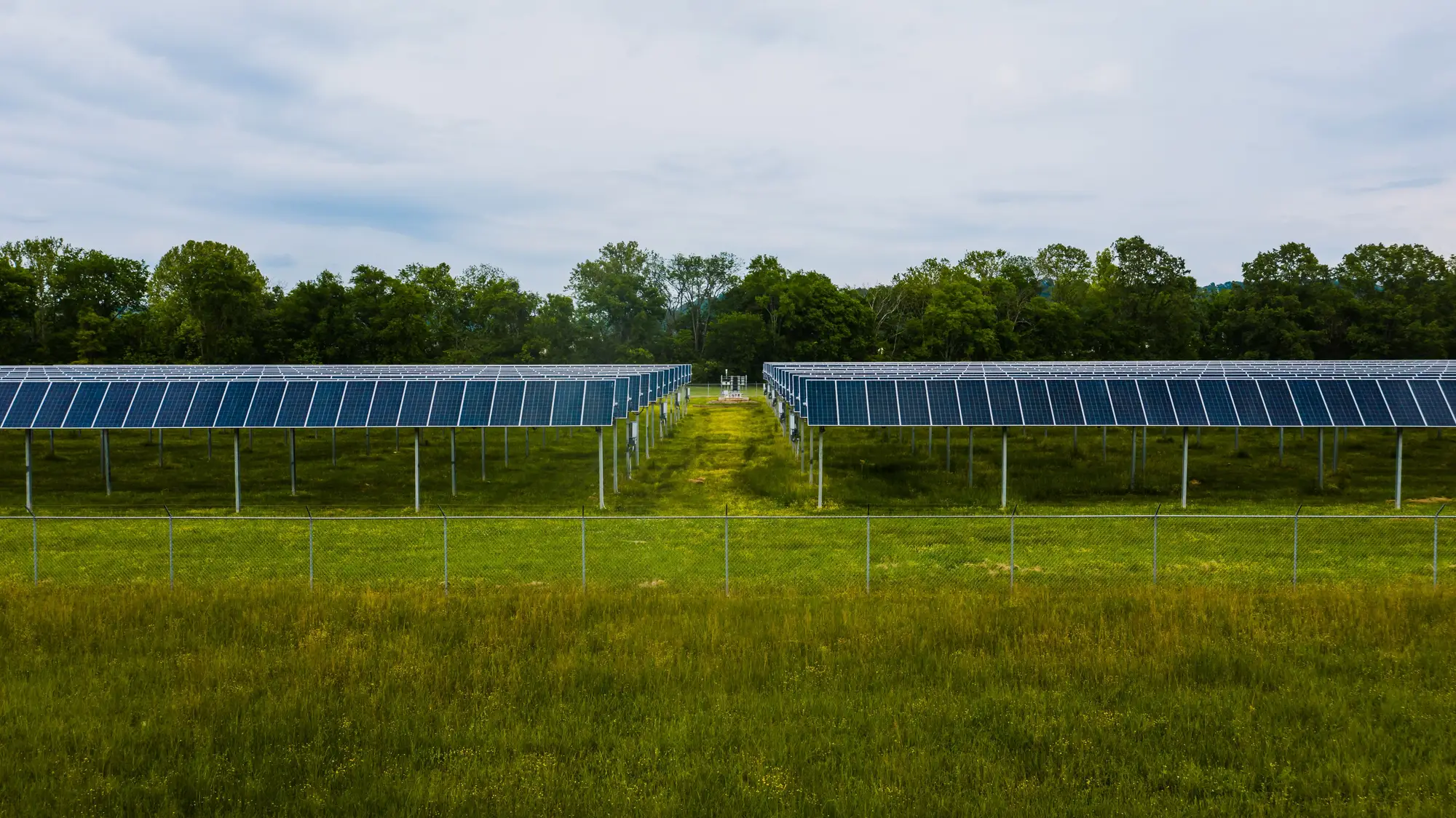 Two rows of ground-mounted solar tables behind a wire fence, seen across a hay field